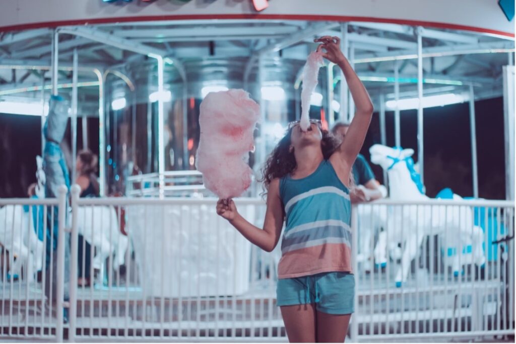 Girl eating cotton candy in front of merry go round. 