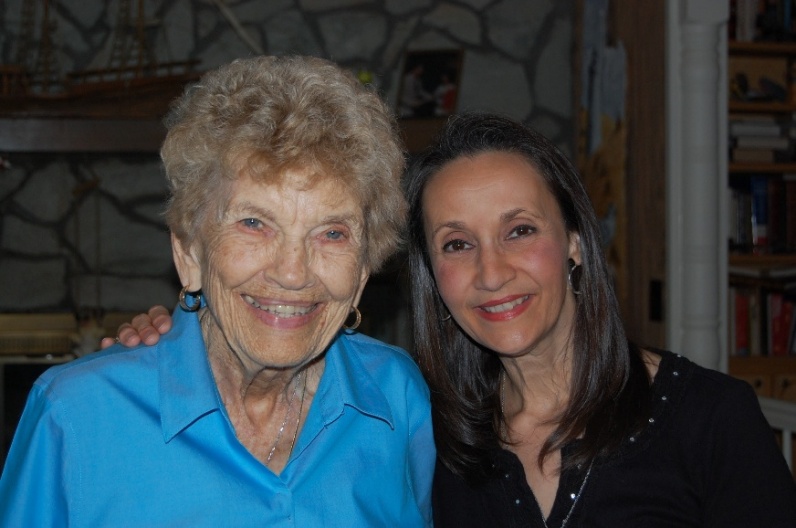 Photograph showing two smiling women posing together indoors. Older woman wears bright blue shirt, while younger woman wears black top; warm, friendly portrait emphasizes their apparent relationship.