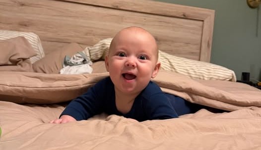 Photograph shows a smiling baby propped up on a bed, facing camera. Warm beige bedding, wooden headboard, pillows, and navy outfit create a cozy bedroom setting.